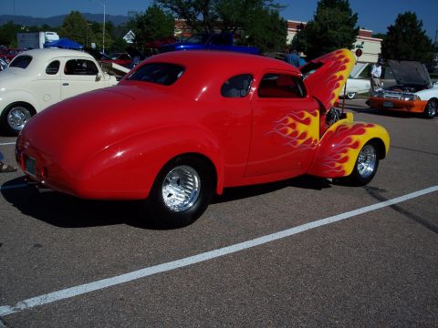 1972 Ford Ranchero - A red hot rod coupe with flame graphics is displayed at a car show, with other vehicles visible in the background.
