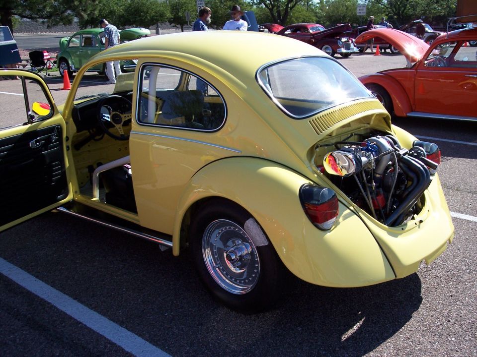 Yellow Volkswagen Beetle, custom engine in rear, roll cage visible inside. Orange VW Beetle and other cars in background