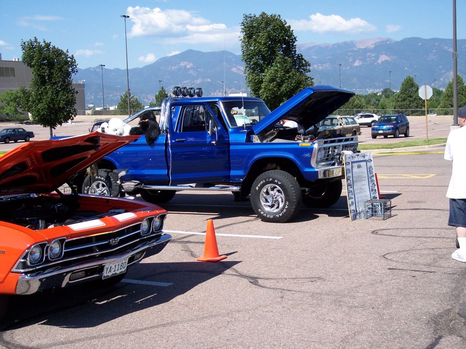 Blue pickup truck with hood open, parked next to an orange muscle car. Mountains in background