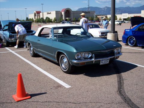 1972 Ford Ranchero - 1965 Chevrolet Corvair convertible at a car show with other vehicles and a mall in the background.