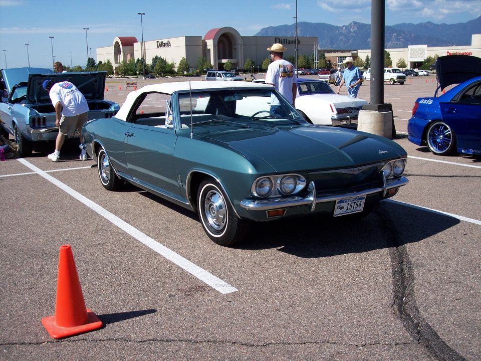 Teal 1965 Chevrolet Corvair convertible parked in a lot with other cars and a shopping mall in background