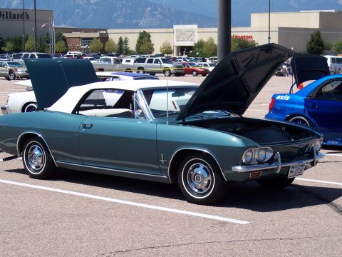 1972 Ford Ranchero - A teal convertible with its hood open, parked in a lot with other vehicles and a shopping mall in the background.