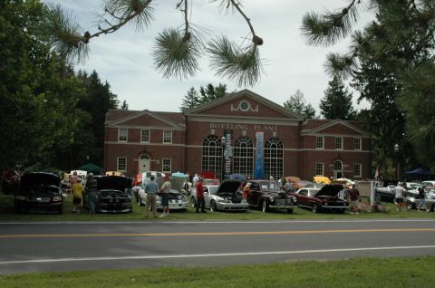 2000 Ford Mustang - A car show is taking place in front of a historic brick building on a cloudy day.