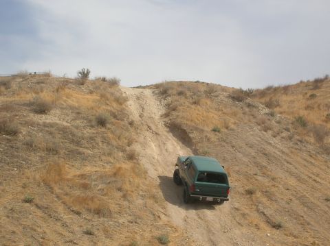 1985 Ford Bronco - A green SUV navigates a steep, dry dirt path under a cloudy sky.