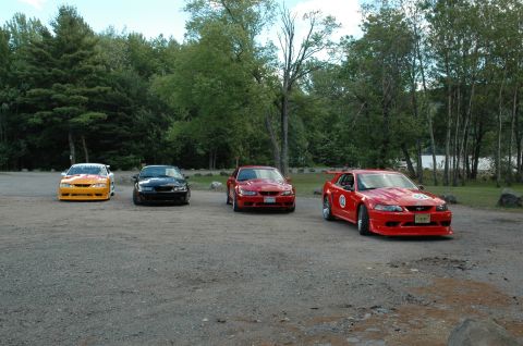 2000 Ford Mustang - Four Ford Mustangs, including a red one with racing decals, are parked in a gravel lot with trees in the background.