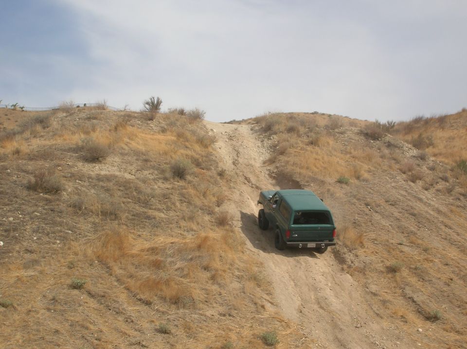 1985 Ford Bronco driving up a steep dirt hill with dry brush and sparse trees