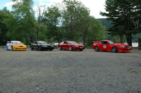 2000 Ford Mustang - A line of four Ford Mustangs, including two red models, a black one, and a custom orange and white variant, are parked on a gravel surface.