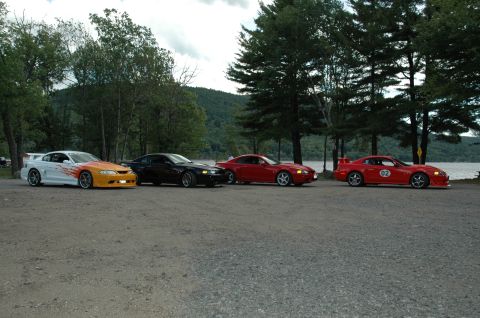2000 Ford Mustang - Four Ford Mustangs are lined up next to a lake, with the red car on the far right displaying a "02" decal.