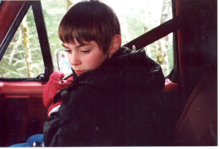 Young boy in a dark winter coat sitting inside a truck cab with red interior, looking down