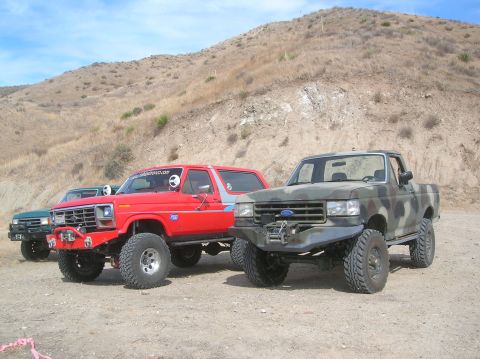 1985 Ford Bronco - Modified Ford Bronco and pickup trucks parked outdoors on a sunny day.
