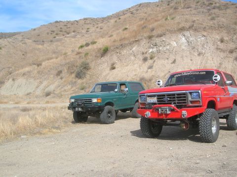 1985 Ford Bronco - Two Ford Broncos, one red and one green, are parked off-road on a dirt path with a barren hillside in the background.