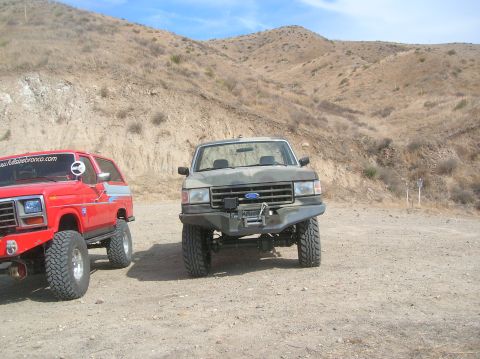 1985 Ford Bronco - Two off-road vehicles are parked on a dirt lot with hills in the background.