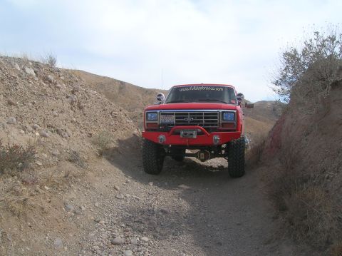 1985 Ford Bronco - Modified red Ford Bronco driving on a rocky, off-road trail under a partly cloudy sky.