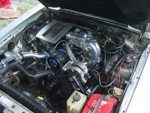 1986 Ford Mustang - Engine bay of a 1986 Ford Mustang, showing a modified engine with aftermarket parts and a red battery.