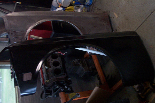 Primered and glossy black fenders leaning in a garage with a bare engine block behind