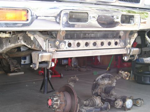 1986 Toyota Hilux - Front underside of a truck on jack stands with the axle pulled, showing a chrome bumper, amber turn signal lens and a bare-metal crossmembe…
