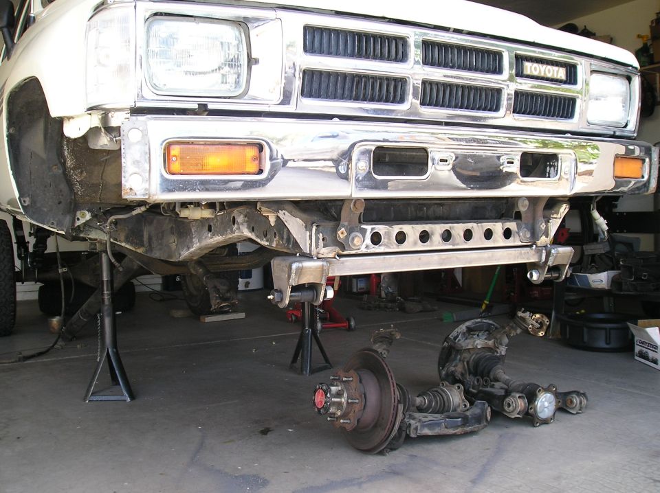 White Toyota truck front end on jack stands, grille and bumper removed, suspension components on the ground
