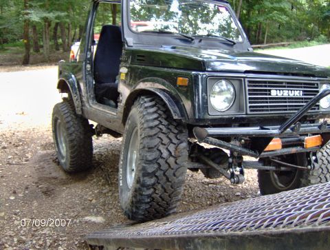 1978 Ford Bronco - A black Suzuki Samurai is positioned on a ramp, its off-road tires prominent against a wooded background.