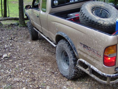 1978 Ford Bronco - A tan pickup truck with off-road tires and a spare tire in the bed is parked on a gravelly surface with trees in the background.