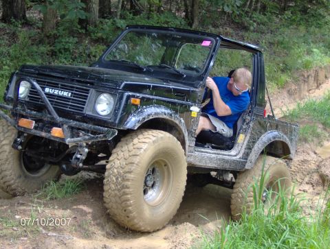 1978 Ford Bronco - A black Suzuki Samurai navigates a muddy off-road course with a driver at the wheel.