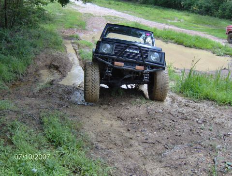 1978 Ford Bronco - A black Suzuki Samurai tackles a challenging muddy off-road course, with another vehicle partially visible in the distance.
