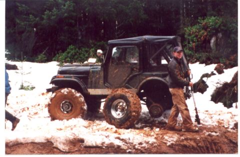 1989 Ford Bronco - Man with rifle stands next to a muddy Jeep CJ-7 in a snowy, wooded area.