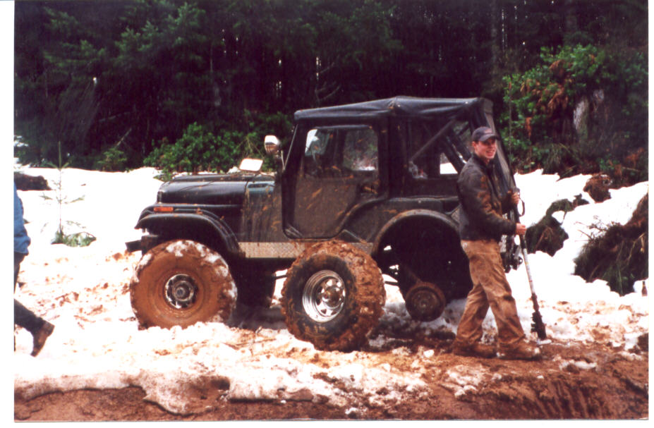 Dark soft-top Jeep CJ on muddy oversized tires in snow, man holding a jack beside it