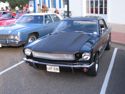 1989 Ford Mustang - A black classic coupe, likely a 1965 Mustang, is parked at a car show next to a blue sedan.