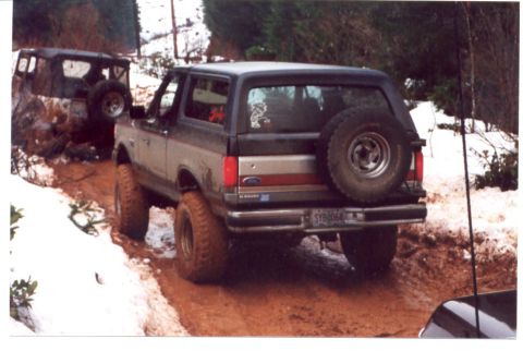 1989 Ford Bronco - A dark gray Ford Bronco navigates a muddy and snowy trail, with another vehicle stuck in the background.