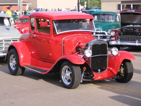 1989 Ford Mustang - A classic red coupe hot rod is displayed at a car show with other vehicles in the background.