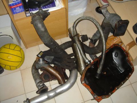 1995 Mazda MX-6 - Various automotive exhaust and oil pan parts are laid out on a tiled floor, with a yellow ball and cabinet visible.