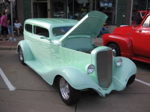 1989 Ford Mustang - A mint green hot rod sedan with flame details is displayed with its hood open, parked beside a red pickup truck.