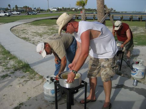 1986 Ford Bronco - Men prepare food outdoors using a propane burner and large pot, with a waterfront view and parked vehicles in the distance.