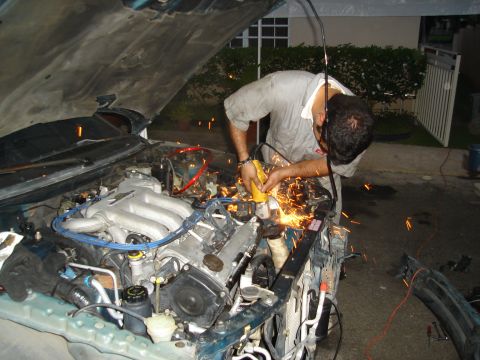 1995 Mazda MX-6 - Mechanic uses a grinder on a car's engine bay, creating a shower of sparks. The vehicle's hood is open.