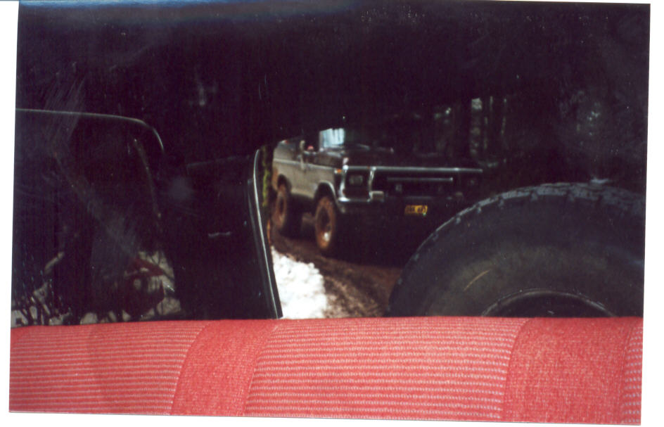 Lifted square-body Ford truck on a muddy snowy trail seen past a red seat and tire