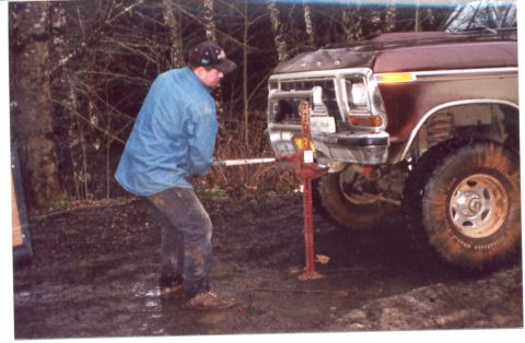 1989 Ford Bronco - Man uses a hi-lift jack to lift the front of a truck in a muddy, wooded area.