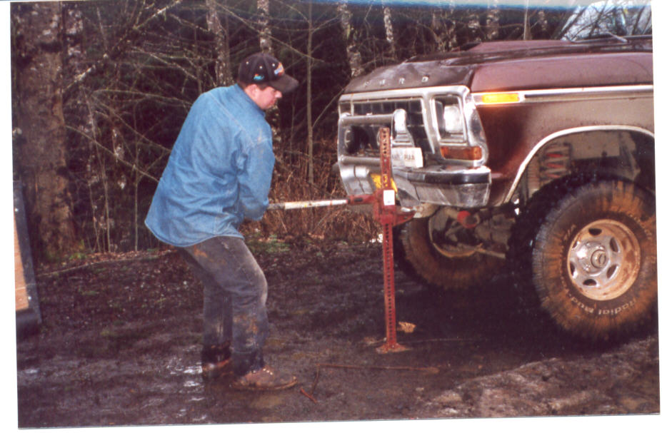 Man using a Hi-Lift jack on the front bumper of a maroon lifted Ford 4x4 in mud