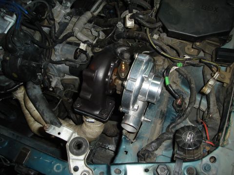 1995 Mazda MX-6 - Close-up view of a turbocharger being installed in a vehicle's engine compartment, with many wires and tubes surrounding it.
