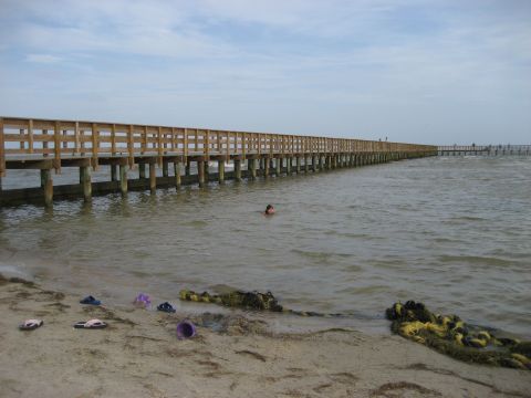1986 Ford Bronco - A long wooden pier stretches over the water, with a swimmer in the distance and flip-flops on the shore.