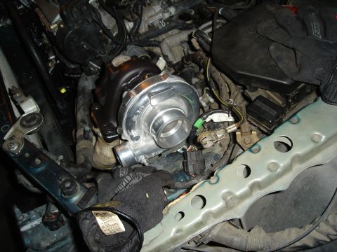 1995 Mazda MX-6 - Close-up view of a turbocharger being installed in a vehicle's engine bay, with hands wearing black gloves visible.