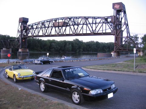 1989 Ford Mustang - A black 1989 Ford Mustang is parked near a bridge, with other classic cars visible.