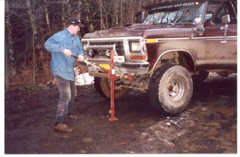 1989 Ford Bronco - A man operates a Hi-Lift jack to lift a vintage Ford truck in a muddy, wooded area.