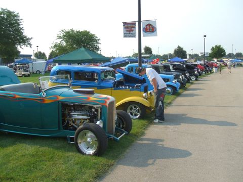 1974 Ford Ranchero - A line of custom cars are displayed at an outdoor event, with spectators milling about.