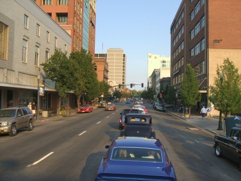 1974 Ford Ranchero - A line of classic cars, including a blue coupe and a black pickup, drive down a city street lined with buildings.