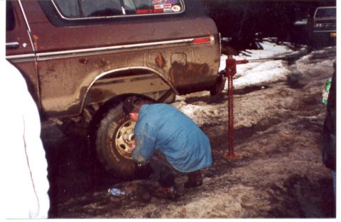 1989 Ford Bronco - A person changes a tire on a brown SUV, which is supported by a red jack. Another vehicle is visible in the background.