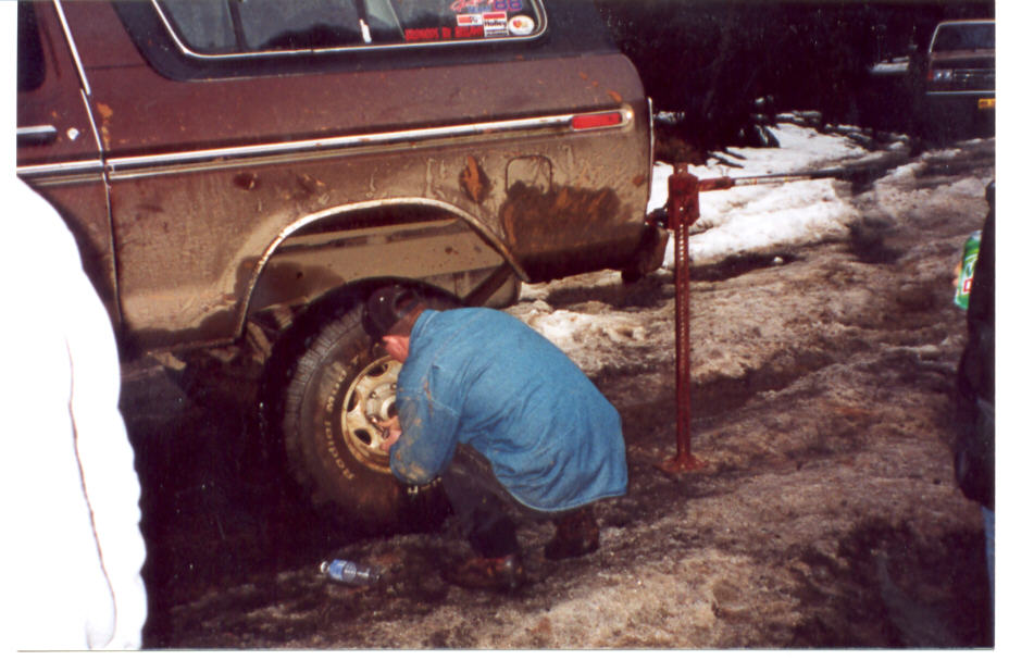 Man changing rear wheel of muddy burgundy Ford Bronco on high-lift jack