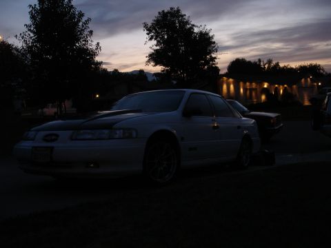 1993 Ford Taurus - A white 1993 Ford Taurus is parked outside as the sun sets, with lights visible in the background.