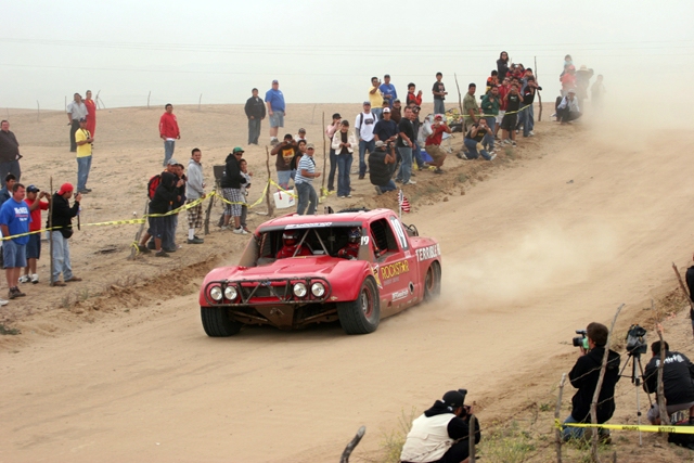 Red off-road race truck with number 19 on door, kicking up dust on a dirt track with spectators watching