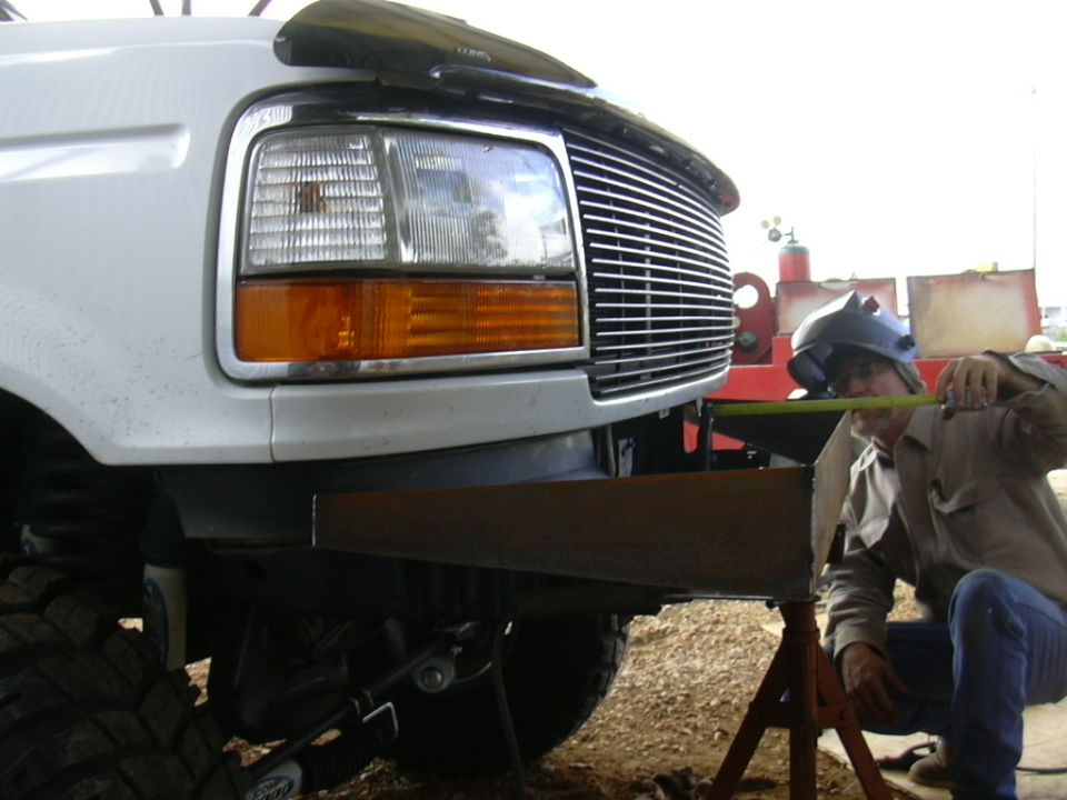 Guy in welding mask measures custom bumper on white Ford truck. Grille and headlights visible
