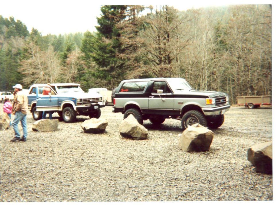 Silver-and-blue lifted Ford Bronco beside a two-tone truck among boulders in a gravel lot